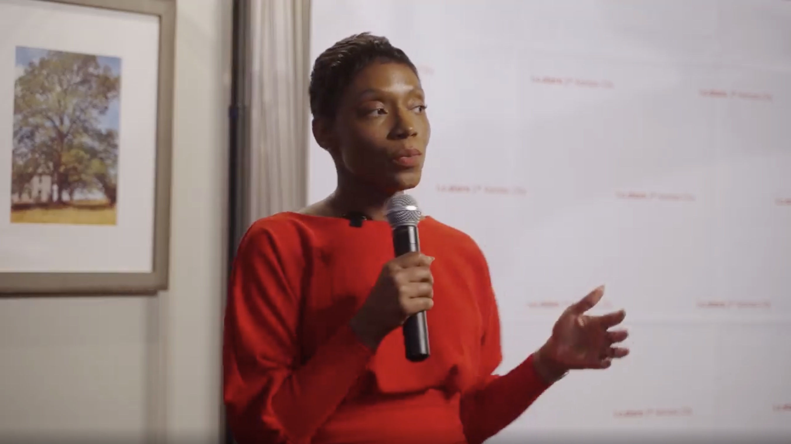 The individual wears a red top, holding a microphone, and appears to be speaking. The background includes a framed painting of a tree and a white backdrop with faint red text, "Vodafone TV 'Serás una diva'."